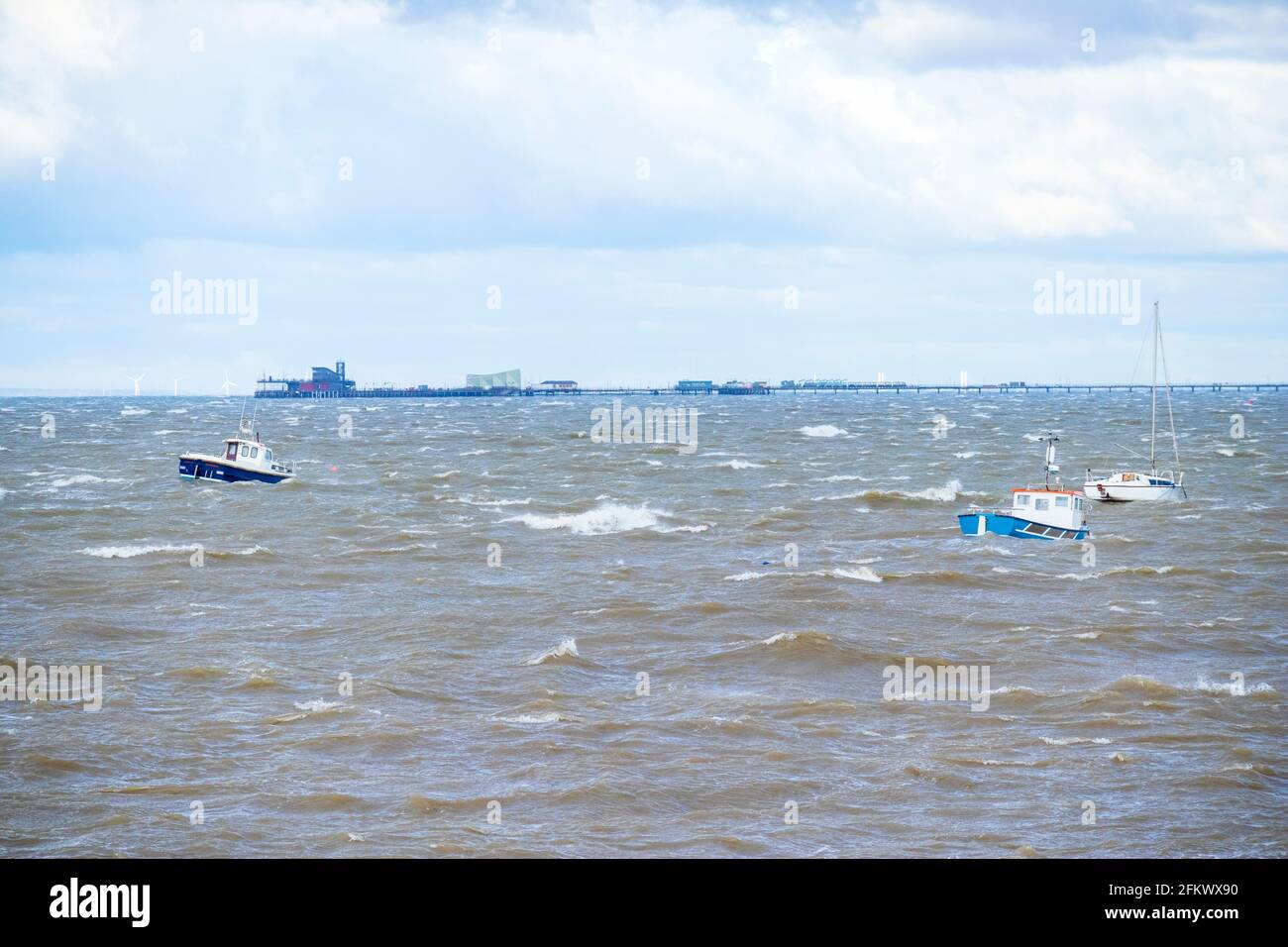 Kleine Boote bei Anchor reiten auf den Wellen bei unsaisonalen Stürmen Anfang Mai an der Themse-Mündung mit Southend Pier Hintergrund Stockfoto