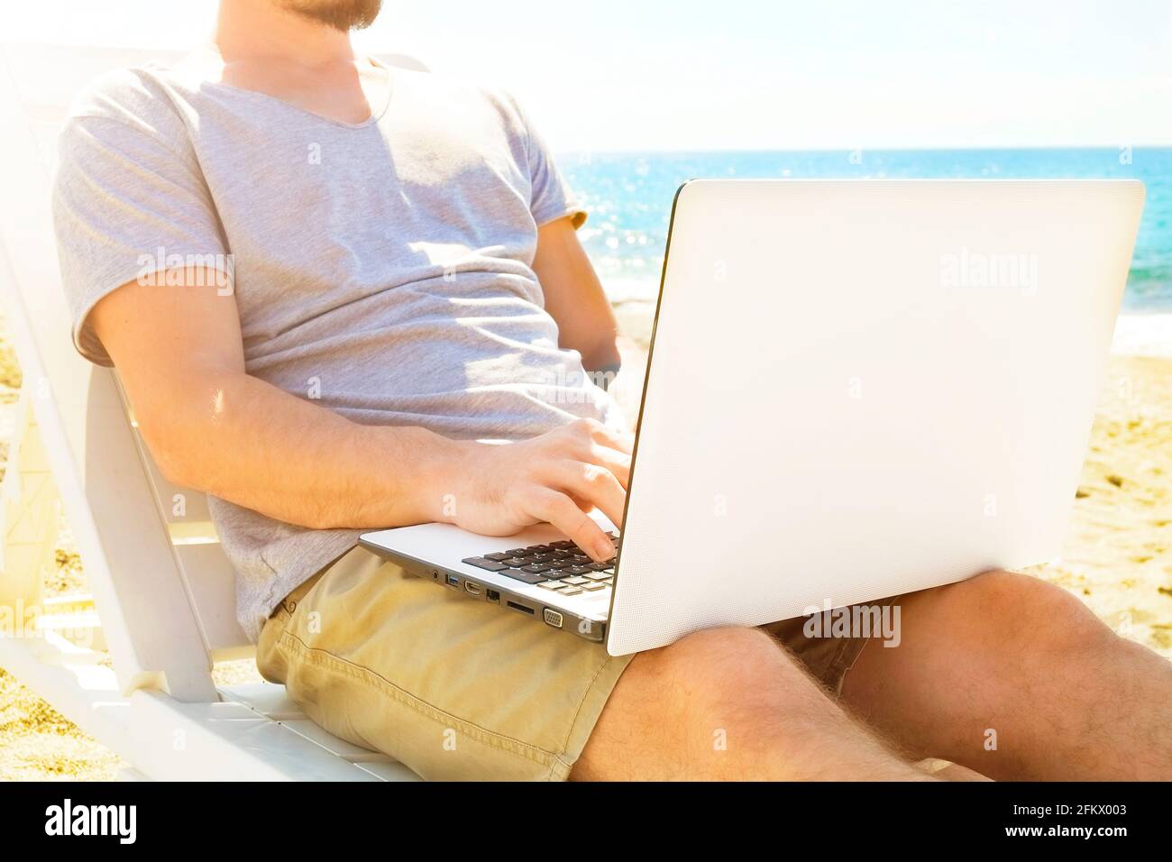 Fit junger Mann sitzt auf Liegestuhl am Strand mit Meerblick mit Laptop. Männlicher freiberuflicher Programmierer in Chaise-Long Lounge, der beim Bloggen beim Surfen arbeitet Stockfoto