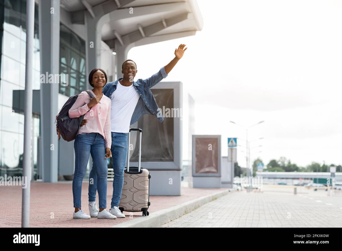 Glückliches Junges Schwarzes Paar, Das Mit Koffer In Der Nähe Des Flughafenterminals Steht Und Ein Taxi Nimmt Stockfoto