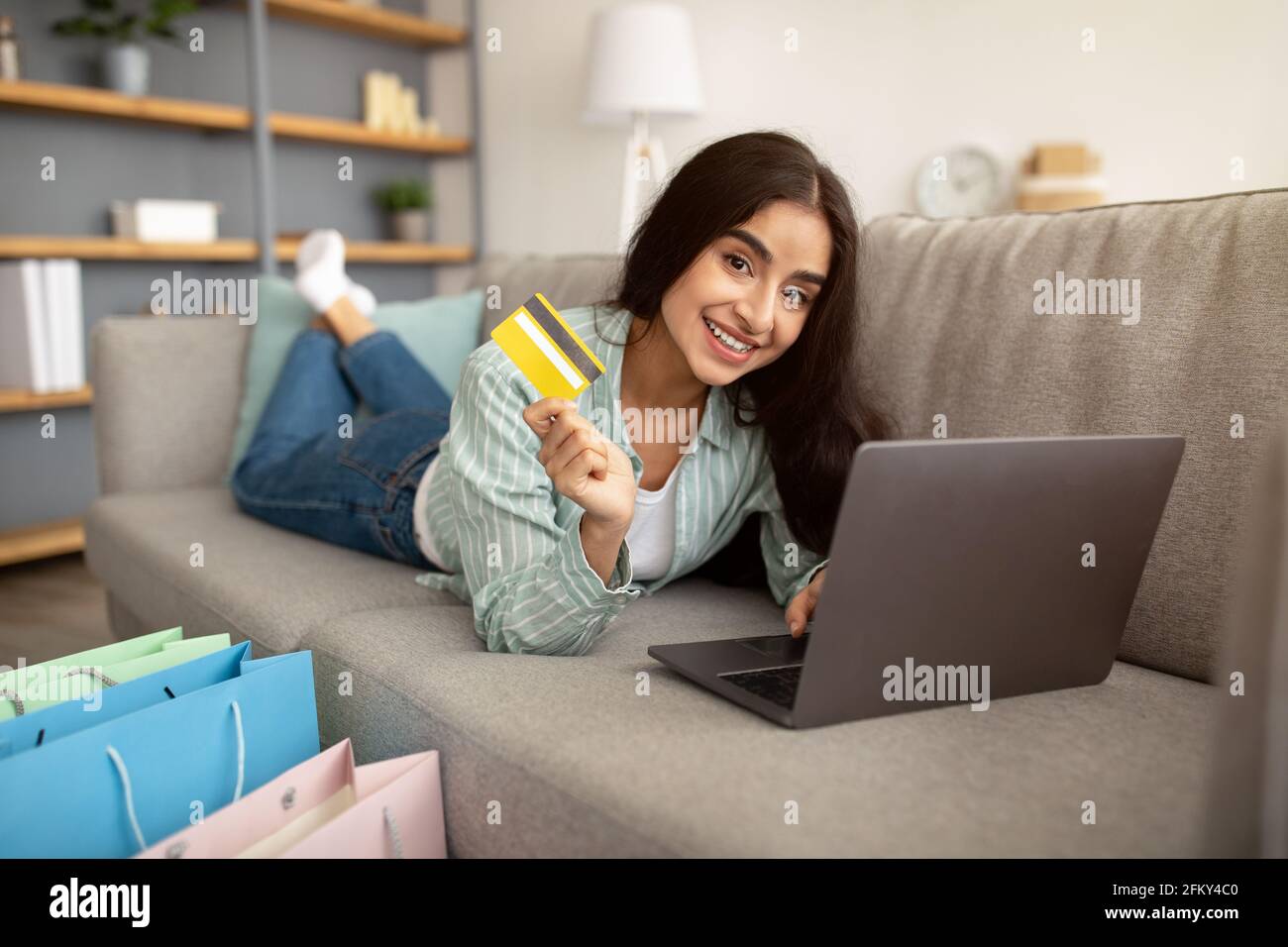 Eine wunderschöne indische Dame, die online einkaufen kann, auf dem Sofa mit Laptop-pc und Kreditkarte liegt, in der Nähe stehen Geschenktüten Stockfoto