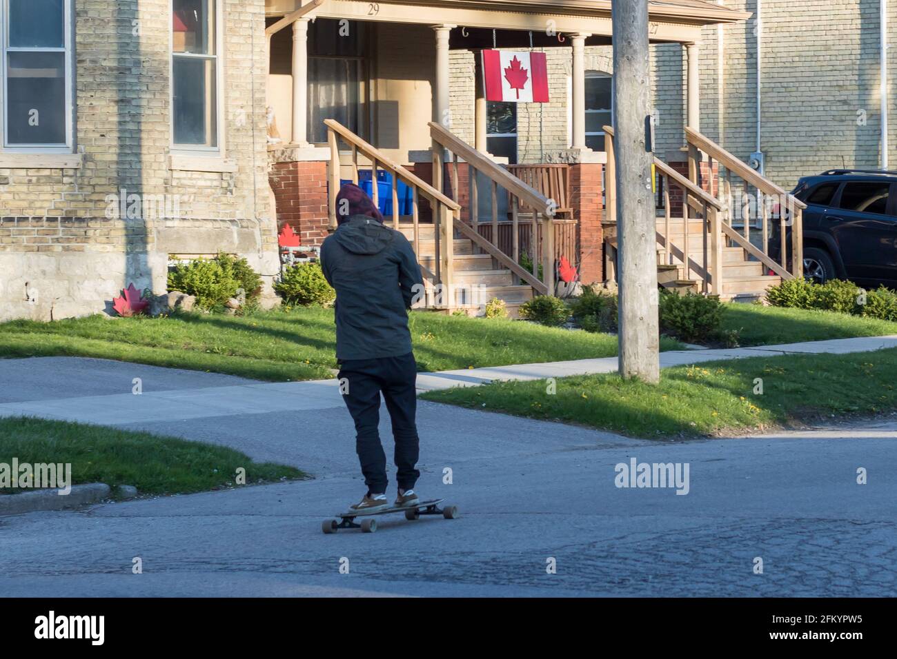 Rückansicht eines jungen Mannes in Jeans, Jacke, Hoodie und Sneakers, der eine Straße entlang in St. Thomas, Ontario, Kanada, Skateboarding macht. Kanada Flagge und Symbole. Stockfoto