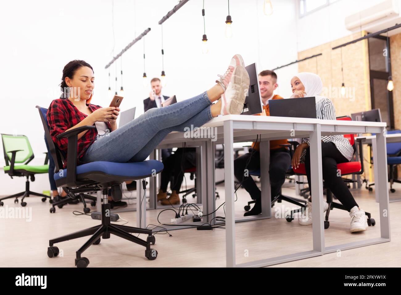 Gruppe von multiethnischen Kollegen, die an Desktop-Computern in einem modernen Bürobereich arbeiten. Mädchen ruht sich mit den Füßen auf dem Tisch aus Stockfoto