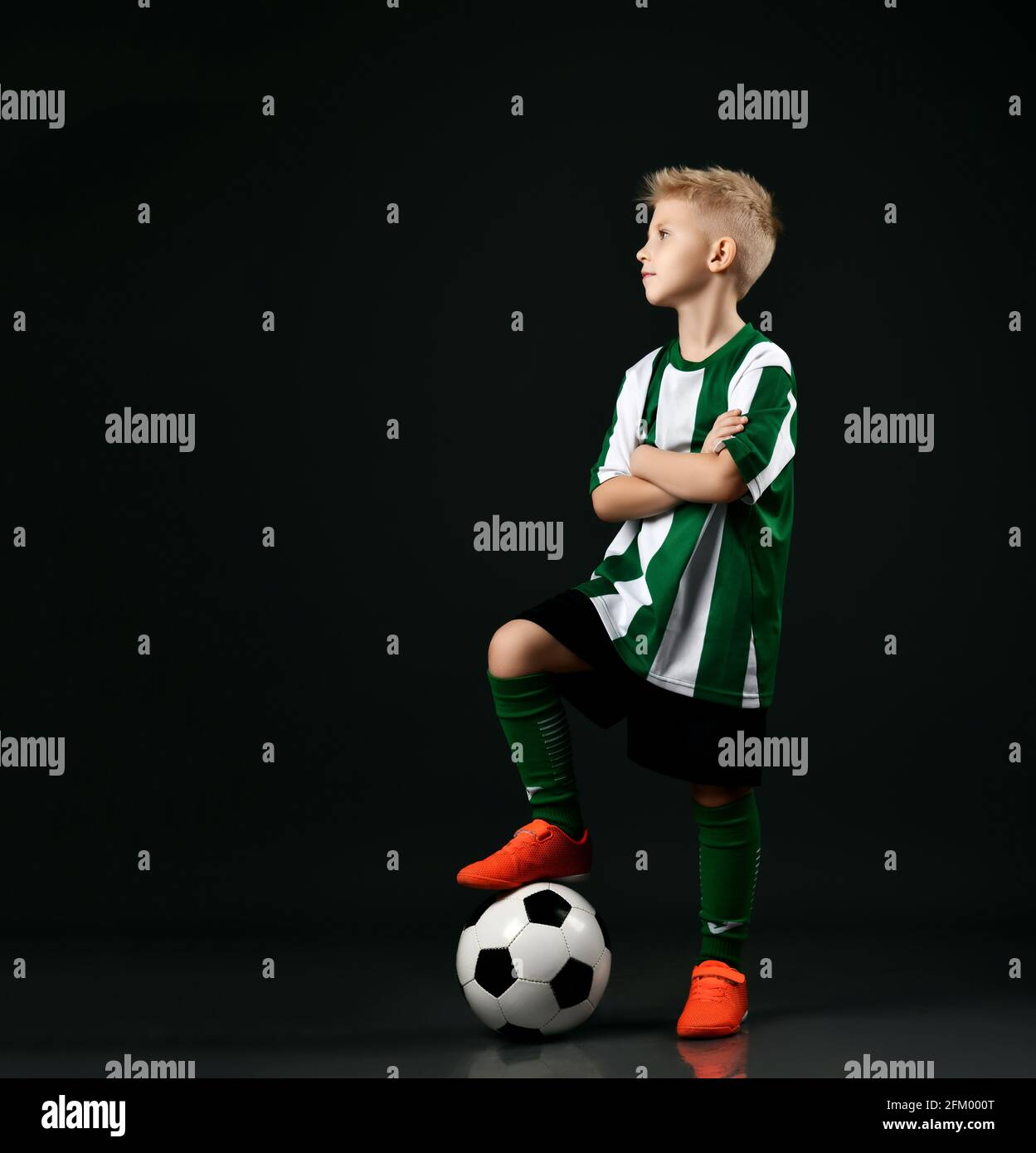 Kleiner Fußballschüler, Junge in rot-weißer Uniform, steht mit Ball unter dem Fuß und schaut zur Seite Stockfoto