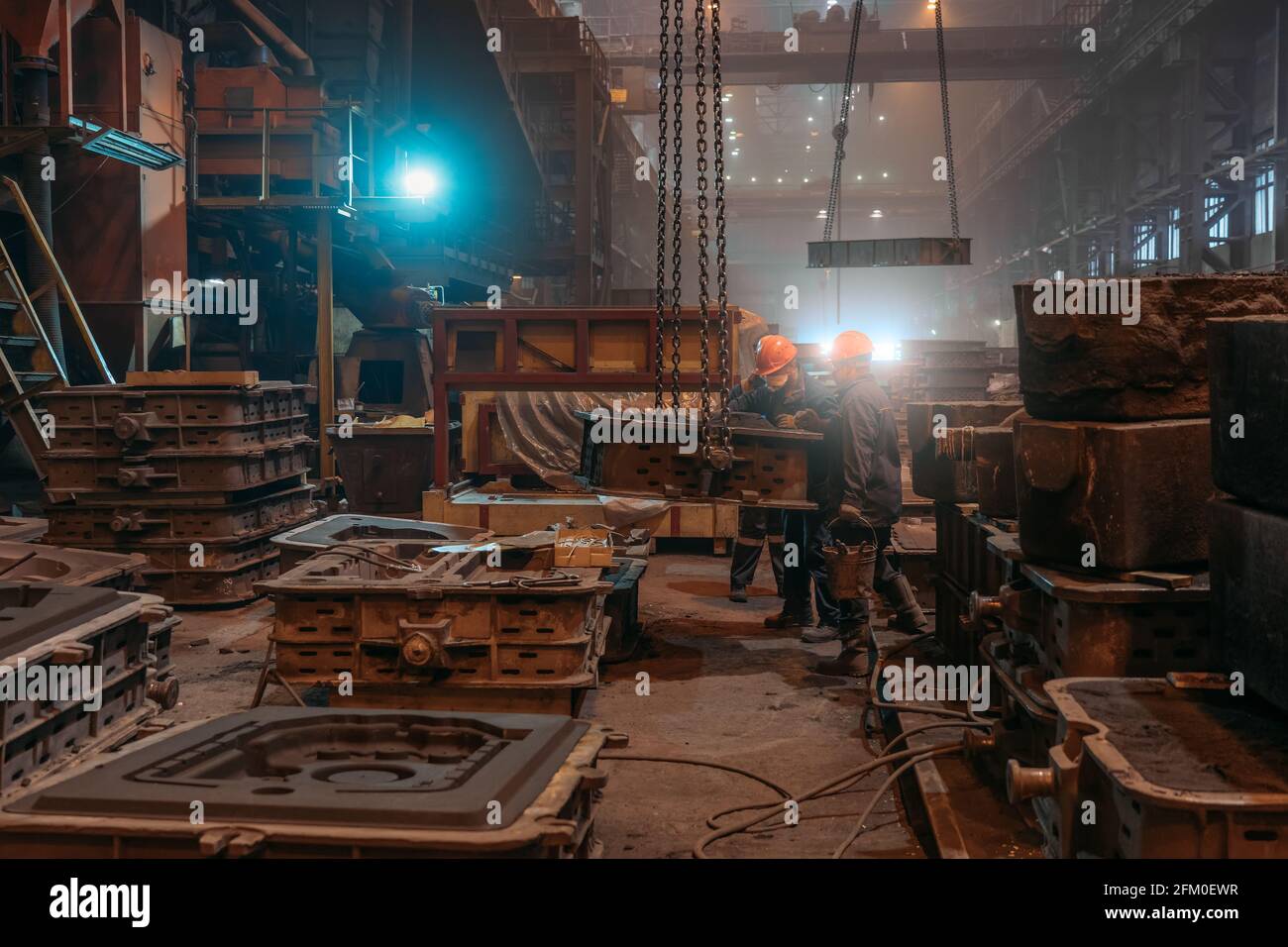 Arbeiter in Helmen und Gesichtsmasken arbeiten nach dem Metallgießen in der Stahlwalzgießerei mit Schimmel an Kranketten. Stockfoto