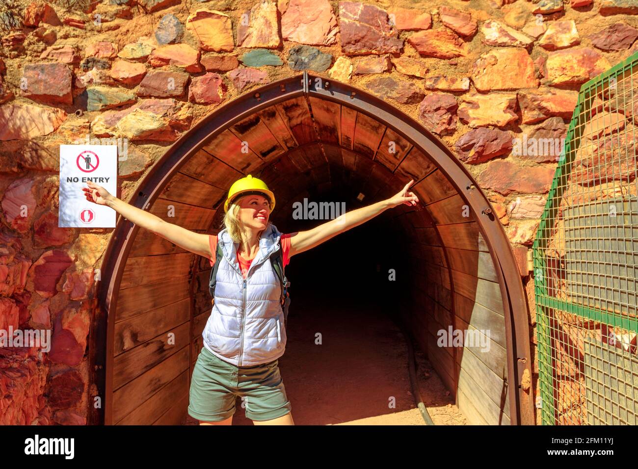 Glückliche kaukasische Touristenfrau mit Bergarbeiterhelm am Eingang der Tunnelhöhle des Battery Hill Mining Center. Tourismus in Tennant Creek im Norden Stockfoto