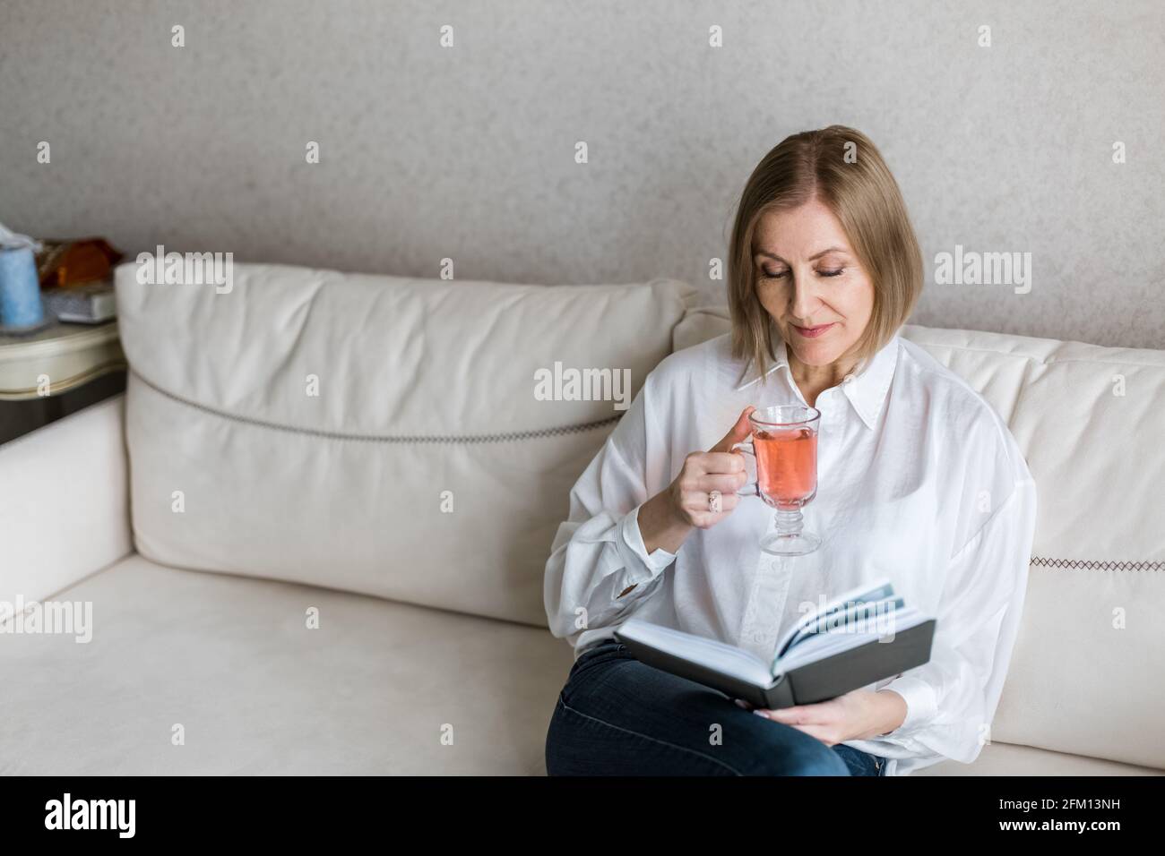 Die Frau sitzt auf der Couch und hält ein Buch in der Hand und trinkt Tee. Stockfoto