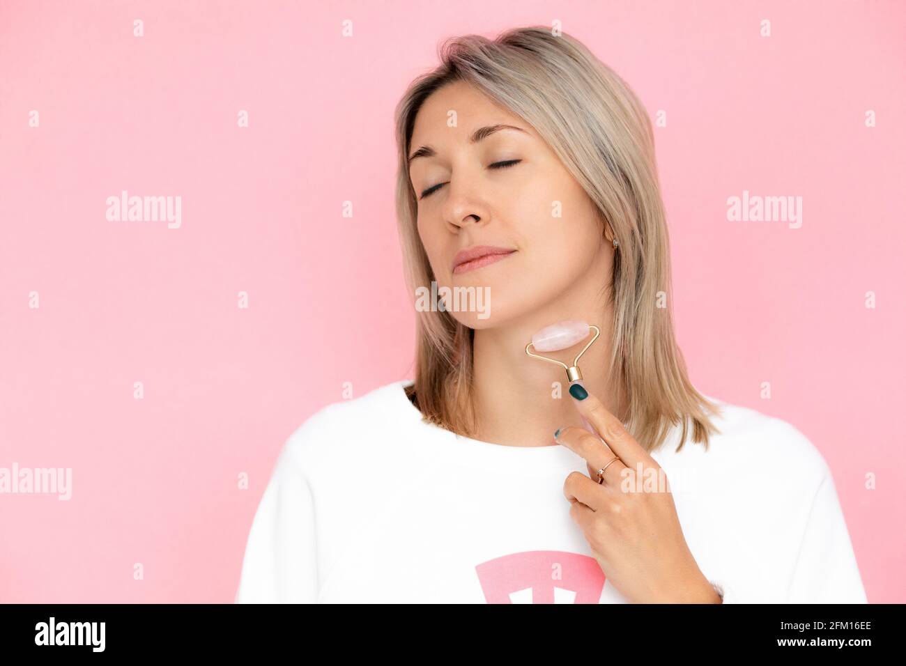 Junge Frau führt Selbstmassage des Halses mit Gua Sha Roller auf rosa Hintergrund. Körperpflegekonzept, Anti-Aging-Pflege, guasha Roller Stockfoto