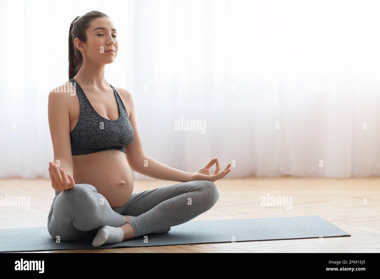 Schöne Schwangerin, die im Wohnzimmer auf einer Yogamatte sitzt und Meditation praktiziert Stockfoto