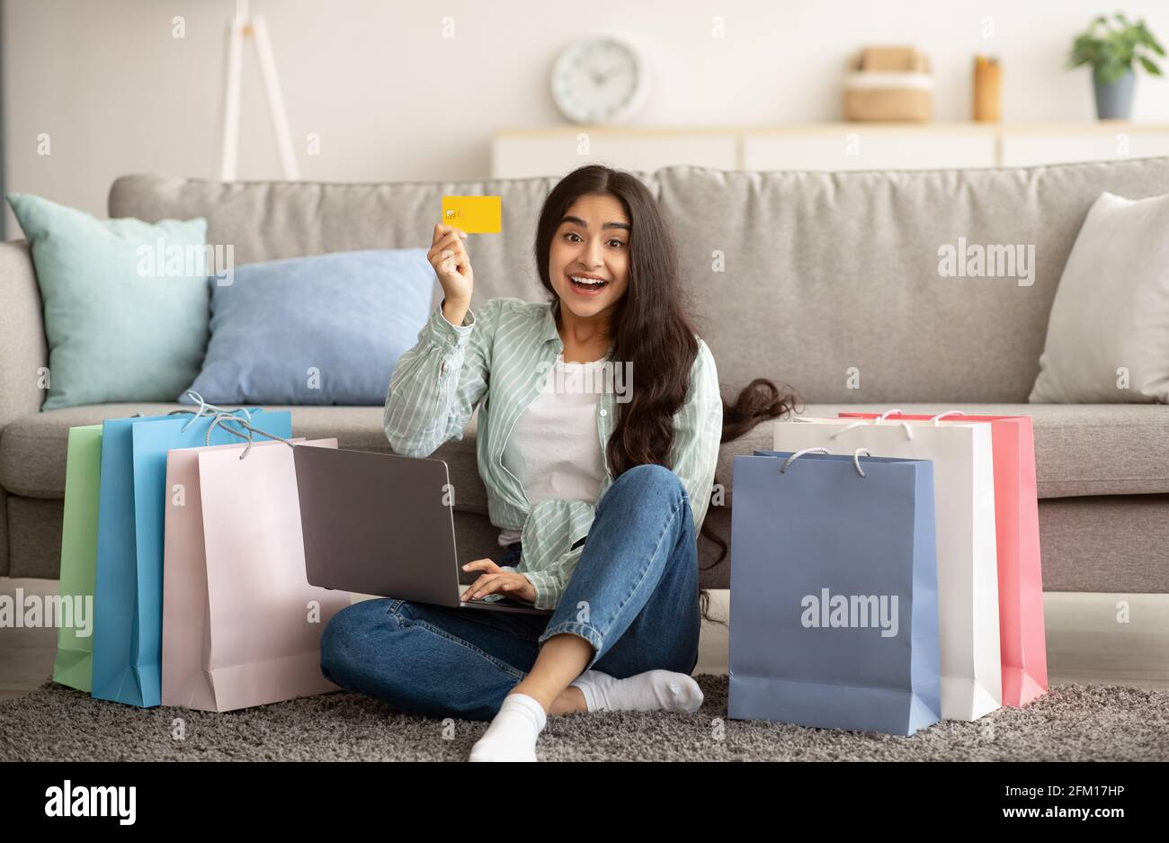 Indische Frau, die sich über den Verkauf im Online-Shop glücklich fühlt, Laptop benutzt, Kreditkarte vorzeigt, mit Geschenktüten auf dem Boden sitzt Stockfoto