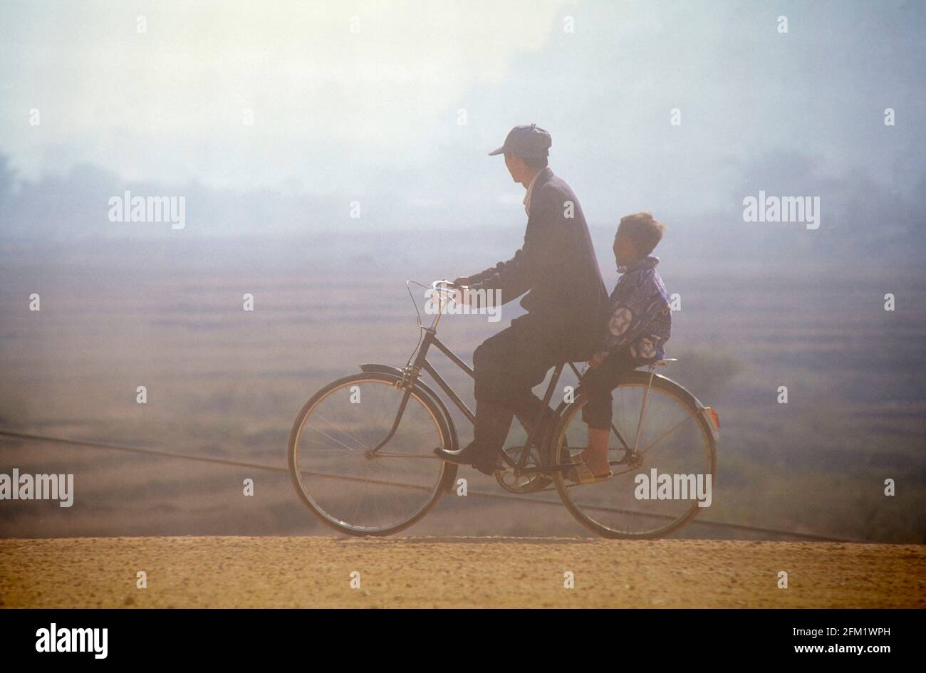 Bei schlichtem Licht fährt ein Vater mit seinem kleinen Sohn auf einem Fahrrad an geernteten Reisfeldern vorbei. 01/1997 - Christoph Keller Stockfoto