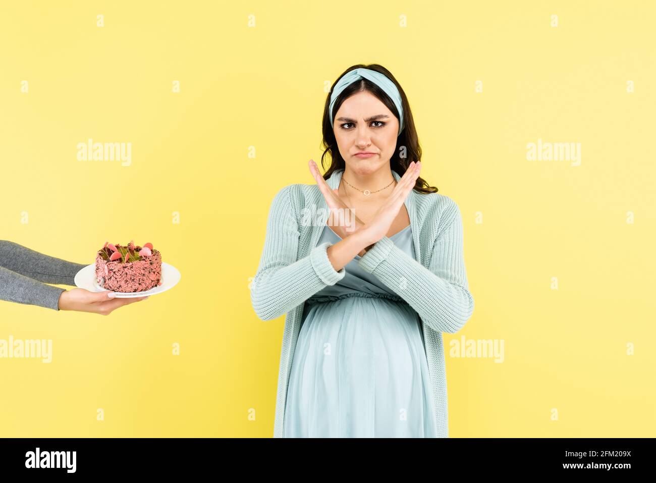 Angewidert, eine Schwangerin zeigt eine abweisende Geste in der Nähe eines auf Gelb isolierten Kuchens Stockfoto