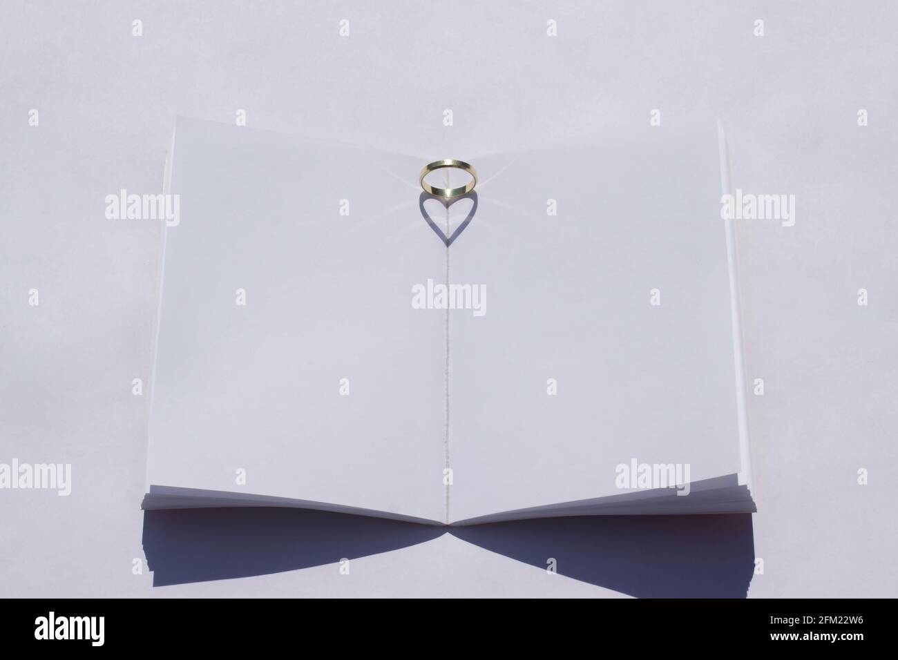 Sommerhochzeit-Mockup. Leeres Hochzeitsbuch auf einem weißen Tisch. Der Ehering wird auf das Buch gelegt und das Sonnenlicht erzeugt einen herzförmigen Schatten. Stockfoto