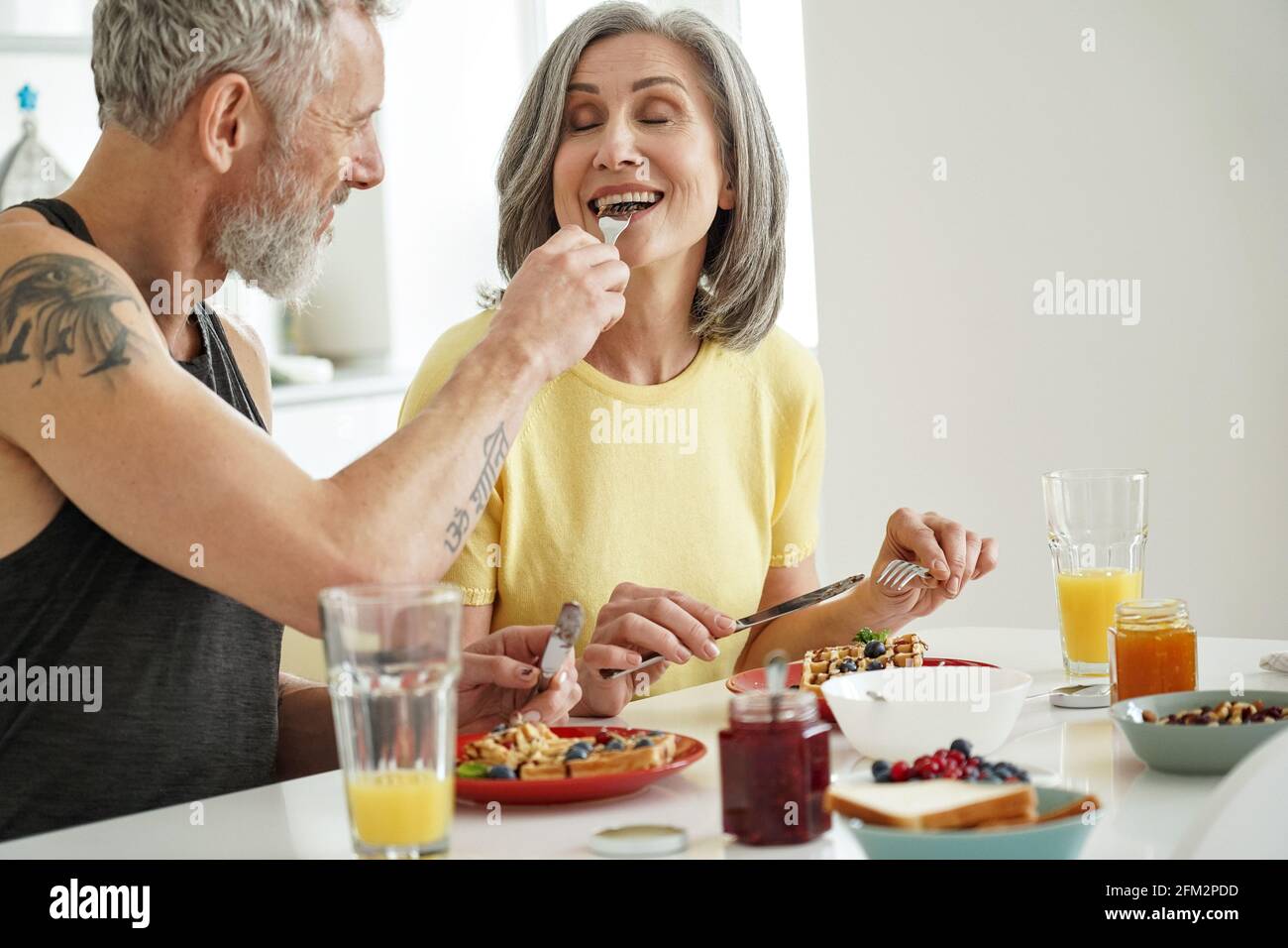 Der ältere Mann füttert seine ältere Frau und probiert Waffeln beim Frühstück zu Hause. Stockfoto