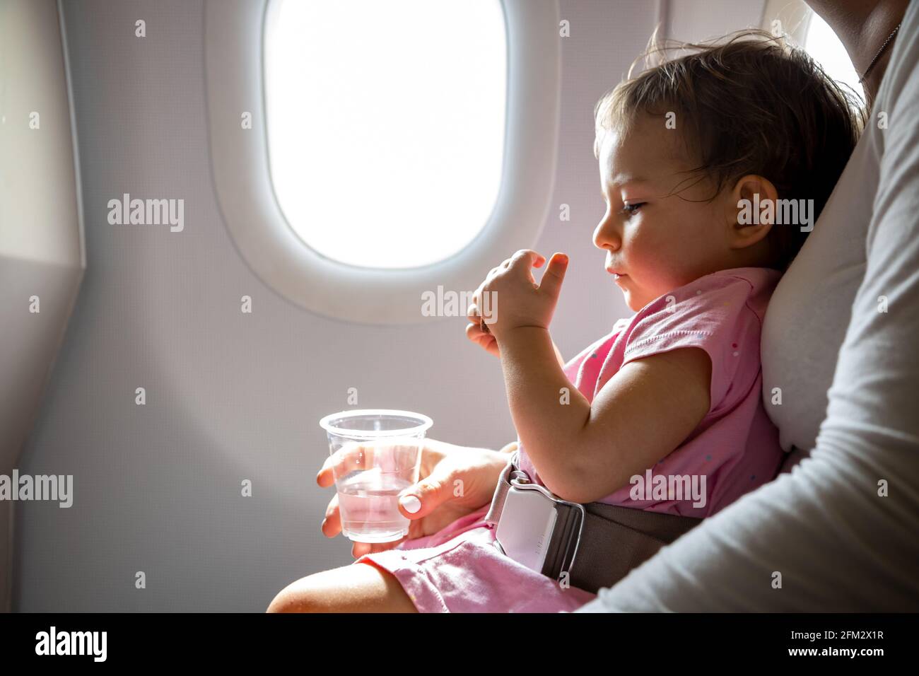 Flug mit Kleinkind. Kleinkind sitzt auf dem Schoß der Mutter, der mit einem speziellen Gürtel im Flugzeug vor dem Bullauge befestigt ist Stockfoto