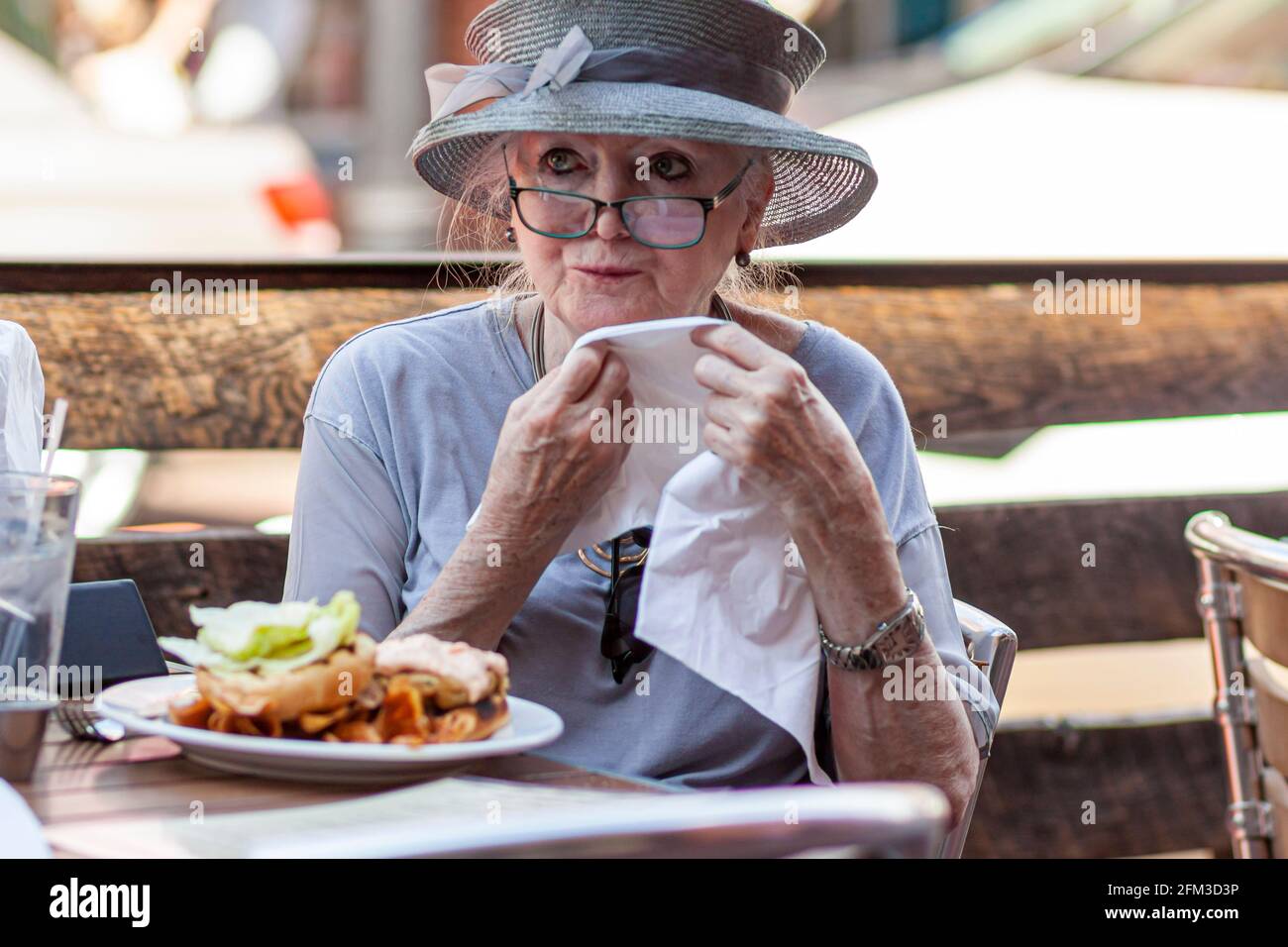 05-02-2021 Annapolis, MD, USA: Eine alte kaukasische Dame mit breitem Hochzeitshut nimmt allein in einem Restaurant zu Mittag. Sie wischt sich den Mund Stockfoto
