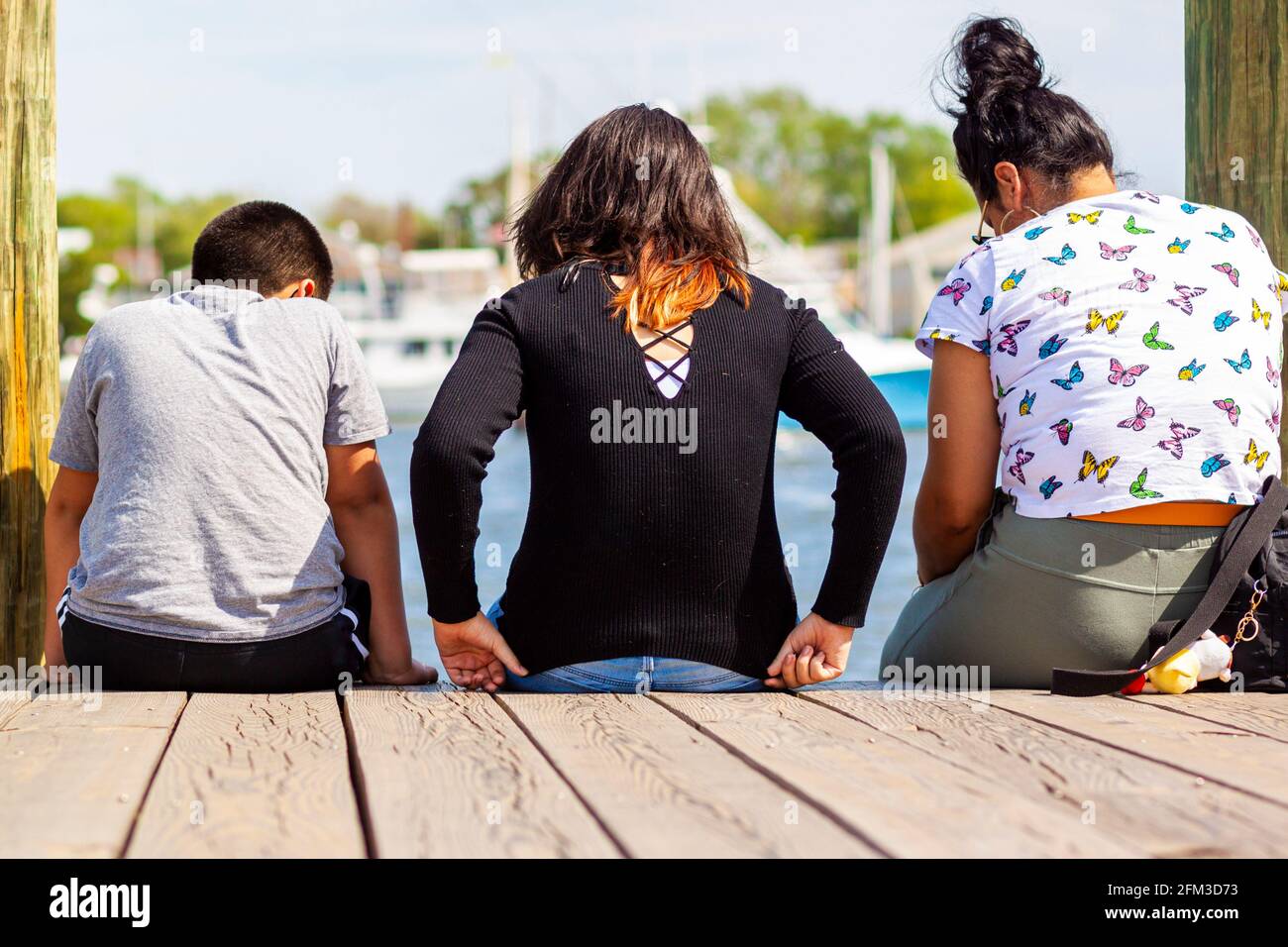 Eine hispanische Familie mit Mutter in der Mitte und ihrem Sohn und ihrer Tochter auf beiden Seiten sitzen auf dem Stadtdock von Annapolis und blicken auf das Meer. Sie Stockfoto