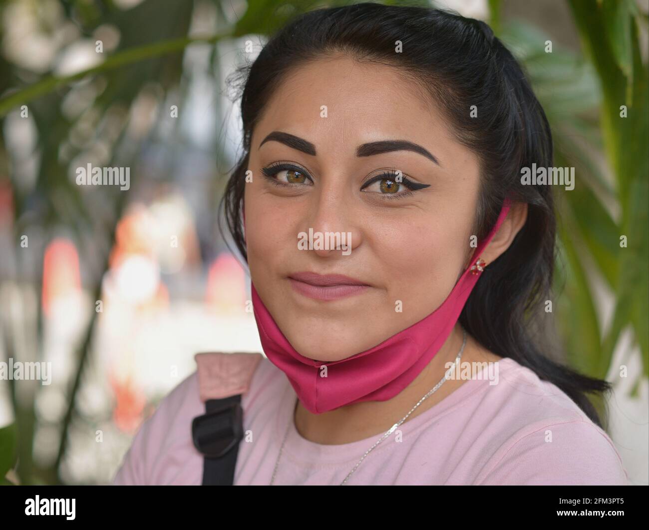 Schöne junge Latina Frau mit modernem Augen Make-up und rosa Gesichtsmaske unter dem Kinn posiert für Kamera und hält soziale Distanz während Corona Pandemie. Stockfoto