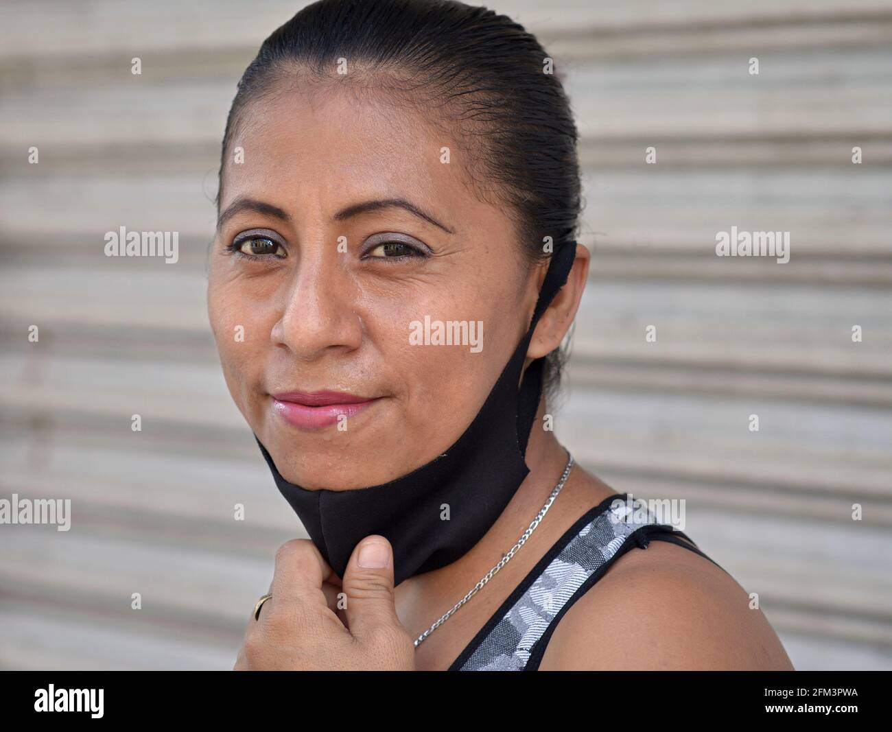 Eine mexikanische Latina-Frau mittleren Alters mit schönen braunen Augen zieht während der globalen Coronavirus-Pandemie eine nicht-medizinische Gesichtsmaske aus schwarzem Stoff nach unten. Stockfoto