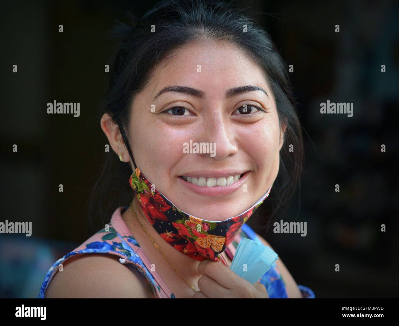 Lächelnde, fröhlich positive junge mexikanische Latina-Frau zieht während der globalen Coronavirus-Pandemie ihre farbenfrohe Gesichtsmaske aus nicht medizinischem Gewebe herunter. Stockfoto