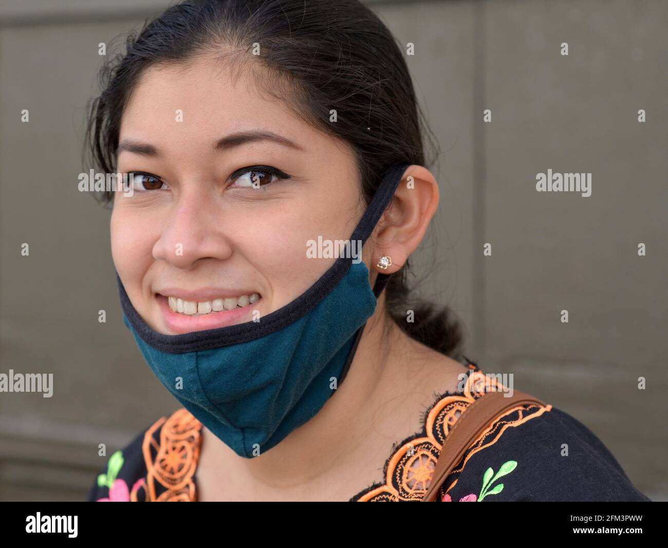 Die charmante, braun-äugige junge Mexikanerin zieht während der globalen Coronavirus-Pandemie ihre blaue Gesichtsmaske aus nicht medizinischem Stoff herunter und lächelt vor der Kamera. Stockfoto