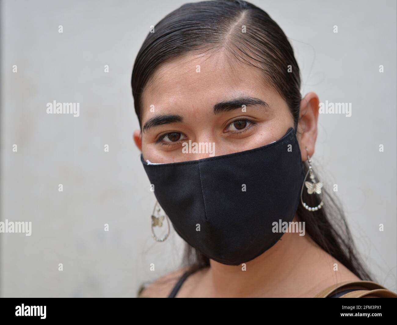 Die junge Latina-Frau mit braunen Augen trägt während der globalen Coronavirus-Pandemie eine schwarze, nicht-medizinische Gesichtsmaske und posiert für die Kamera. Stockfoto