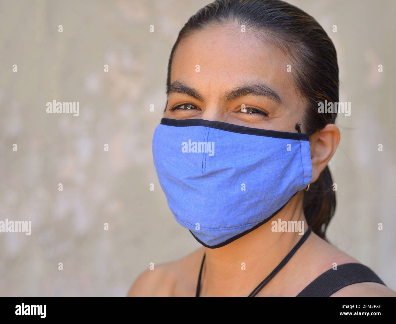 Einfallsreiche mexikanische Latina-Frau mit lächelnden Augen trägt während der globalen Coronavirus-Pandemie eine blaue, nicht-medizinische Gesichtsmaske und posiert für die Kamera. Stockfoto