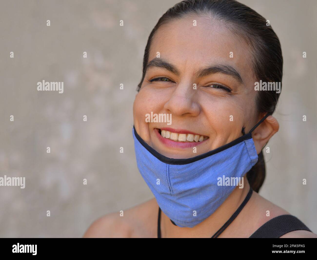 Fröhliche mexikanische Frau mit langen Haaren am Rücken zieht ihre blaue, nicht-medizinische Gesichtsmaske nach unten und lächelt für die Kamera. Stockfoto