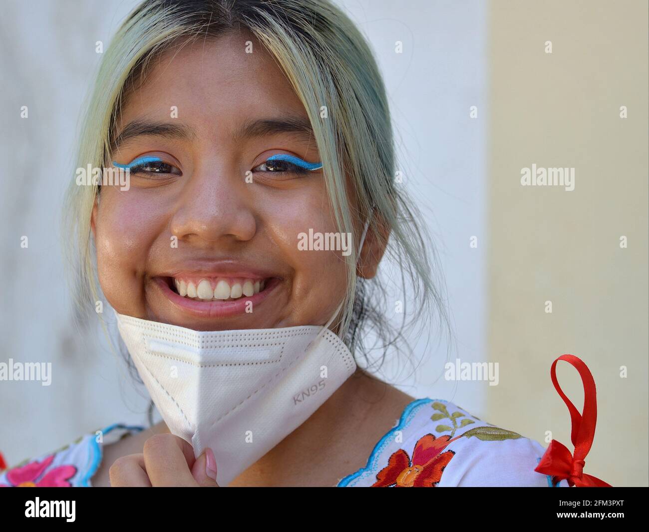 Fröhliches mexikanisches Teenager-Mädchen mit aufwändigem blauem Augen-Make-up zieht ihre weiße KN95-Gesichtsmaske herunter und lächelt während der Coronavirus-Pandemie für die Kamera. Stockfoto