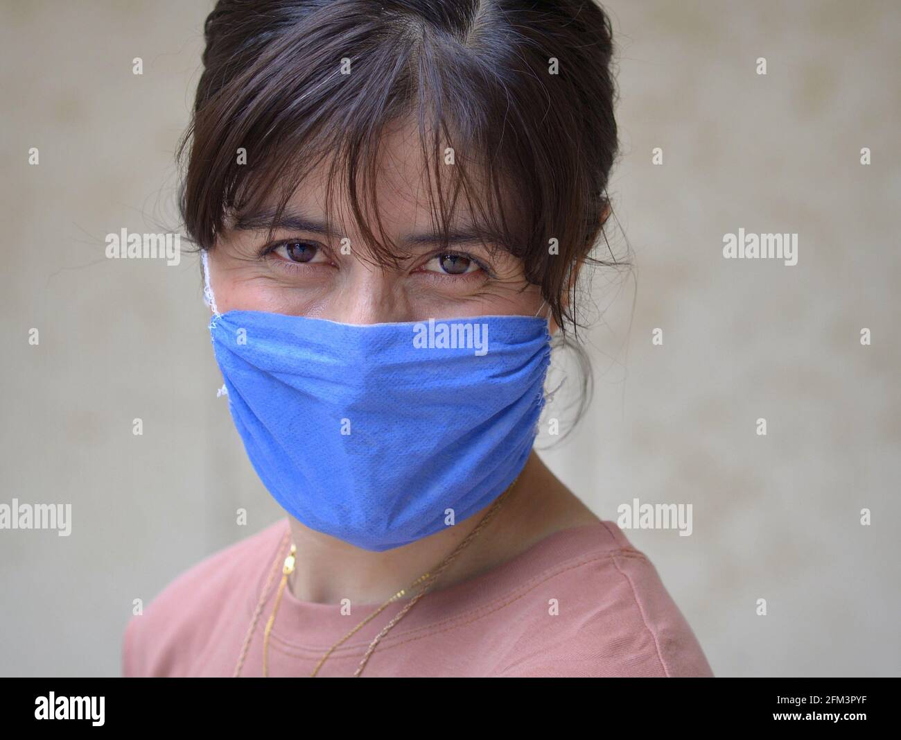 Die junge kaukasische Frau mit lächelnden braunen Augen trägt während der globalen Coronavirus-Pandemie eine selbstgemachte Gesichtsmaske aus nicht-medizinischem blauem Stoff. Stockfoto
