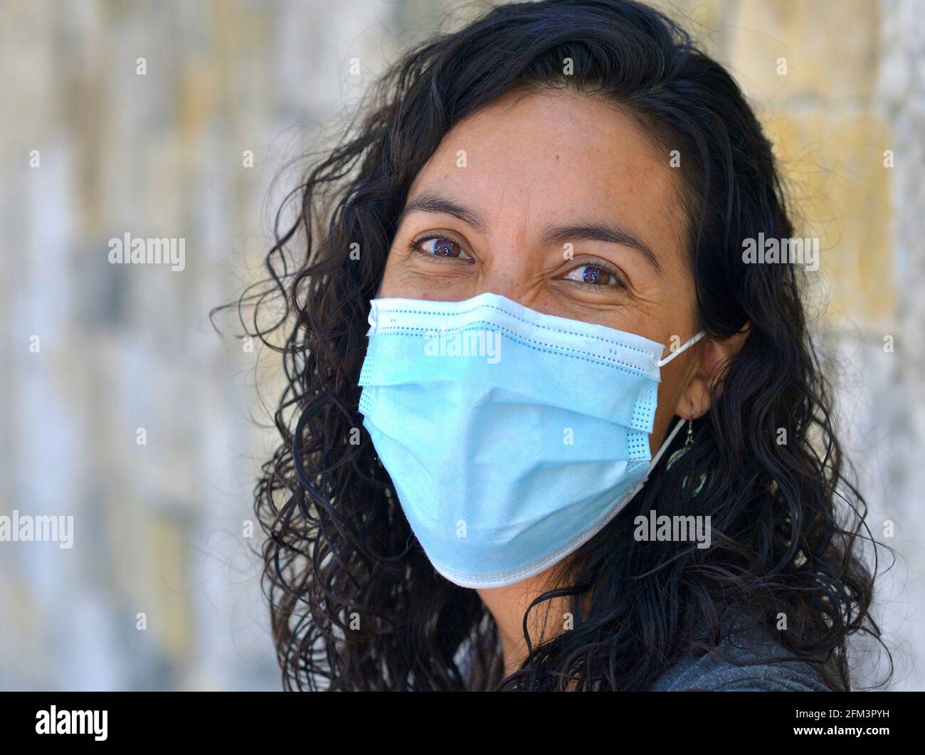 Positive optimistische kaukasische Frau mit nassen langen schwarzen Locken und lächelnden braunen Augen trägt während der Corona-Pandemie eine hellblaue chirurgische Gesichtsmaske. Stockfoto