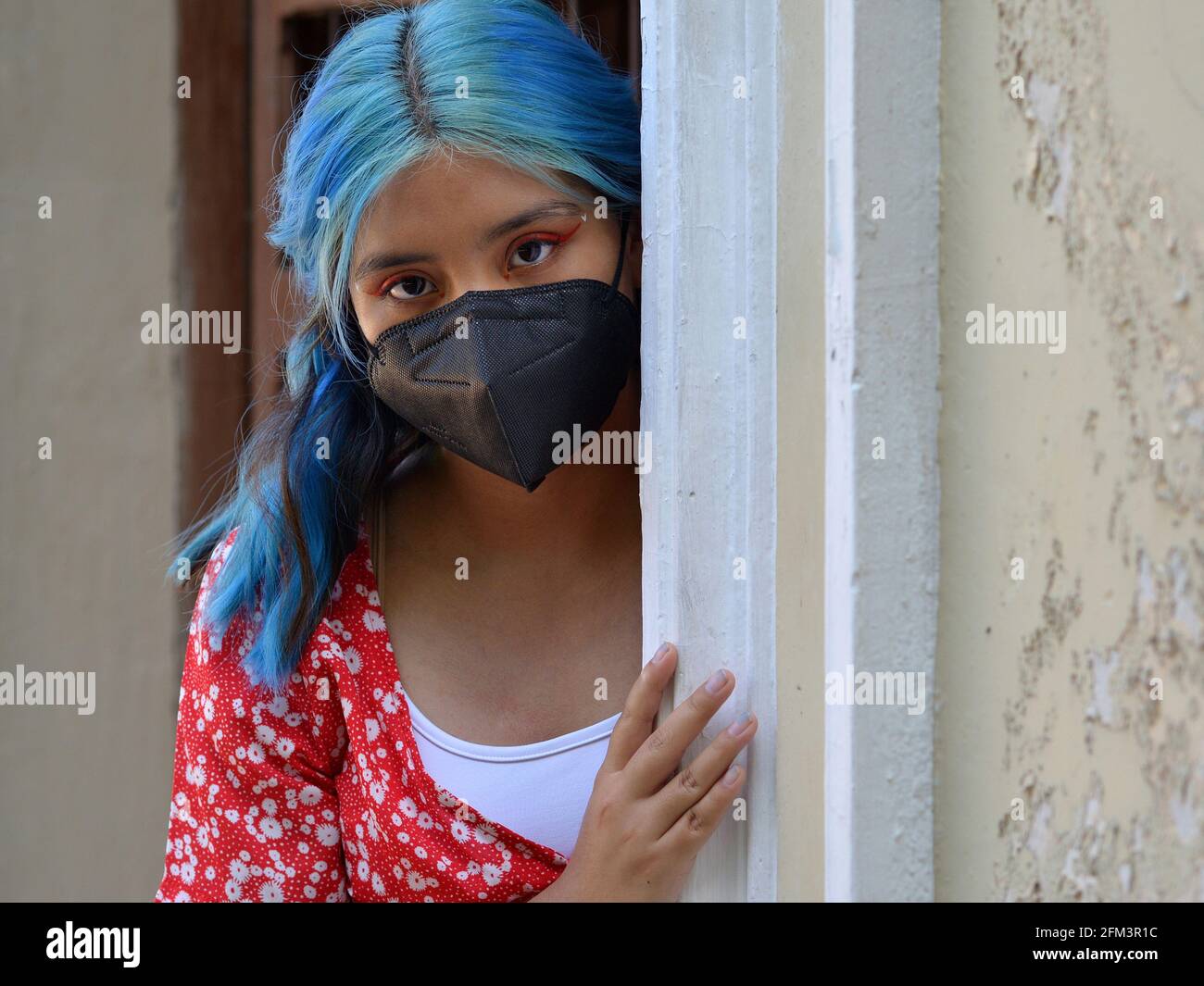 Das kaukasische Latina-Teenager-Mädchen mit blau gefärbtem langen Haar und aufwändigem Augen-Make-up trägt eine schwarze Gesichtsmaske und guckst um die Ecke einer Eingangstür. Stockfoto