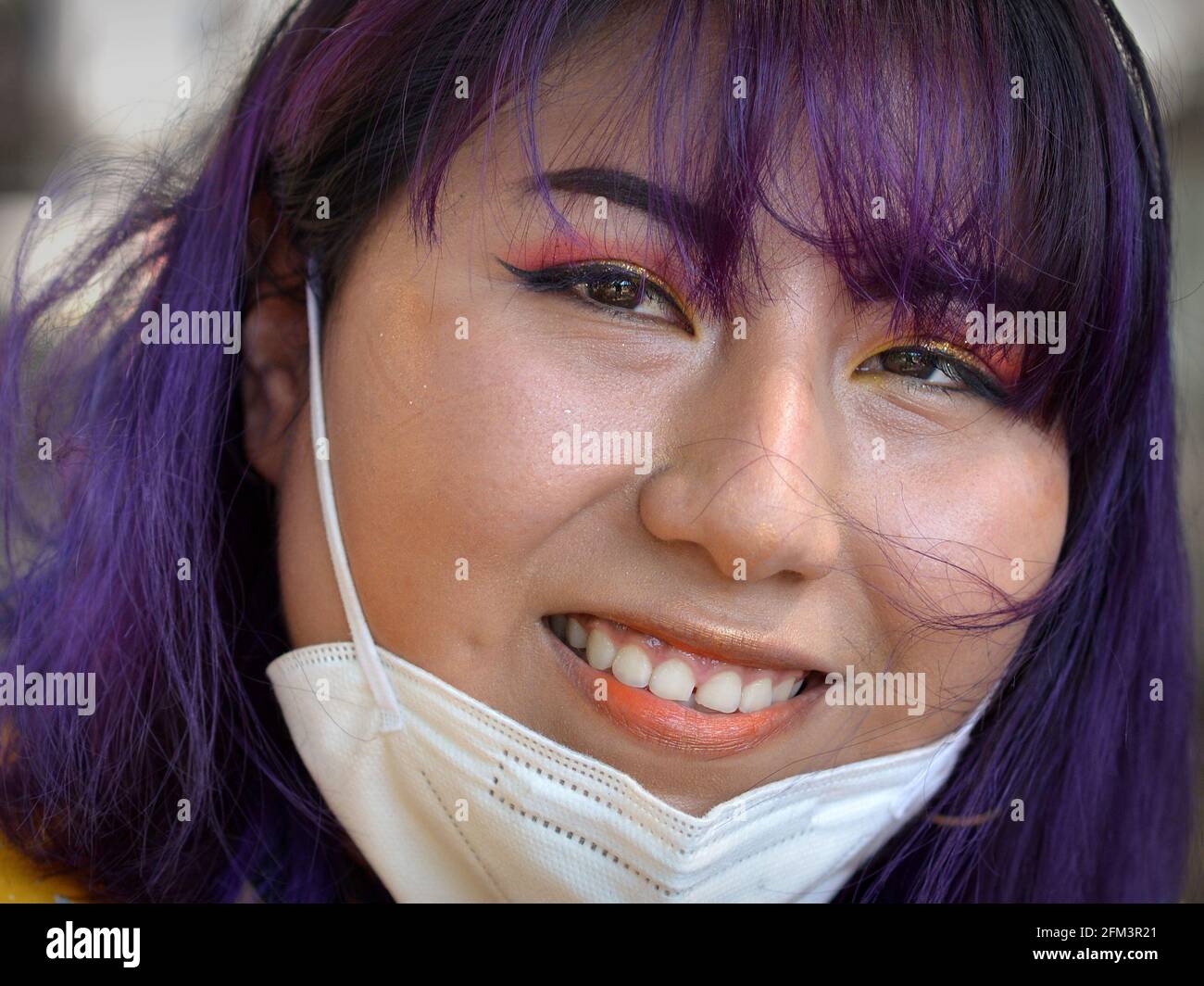 Junge glückliche indigene Südamerikanerin mit aufwändigem Augen-Make-up und blau gefärbtem Haar zieht ihre KN95-Gesichtsmaske herunter und lächelt für die Kamera. Stockfoto