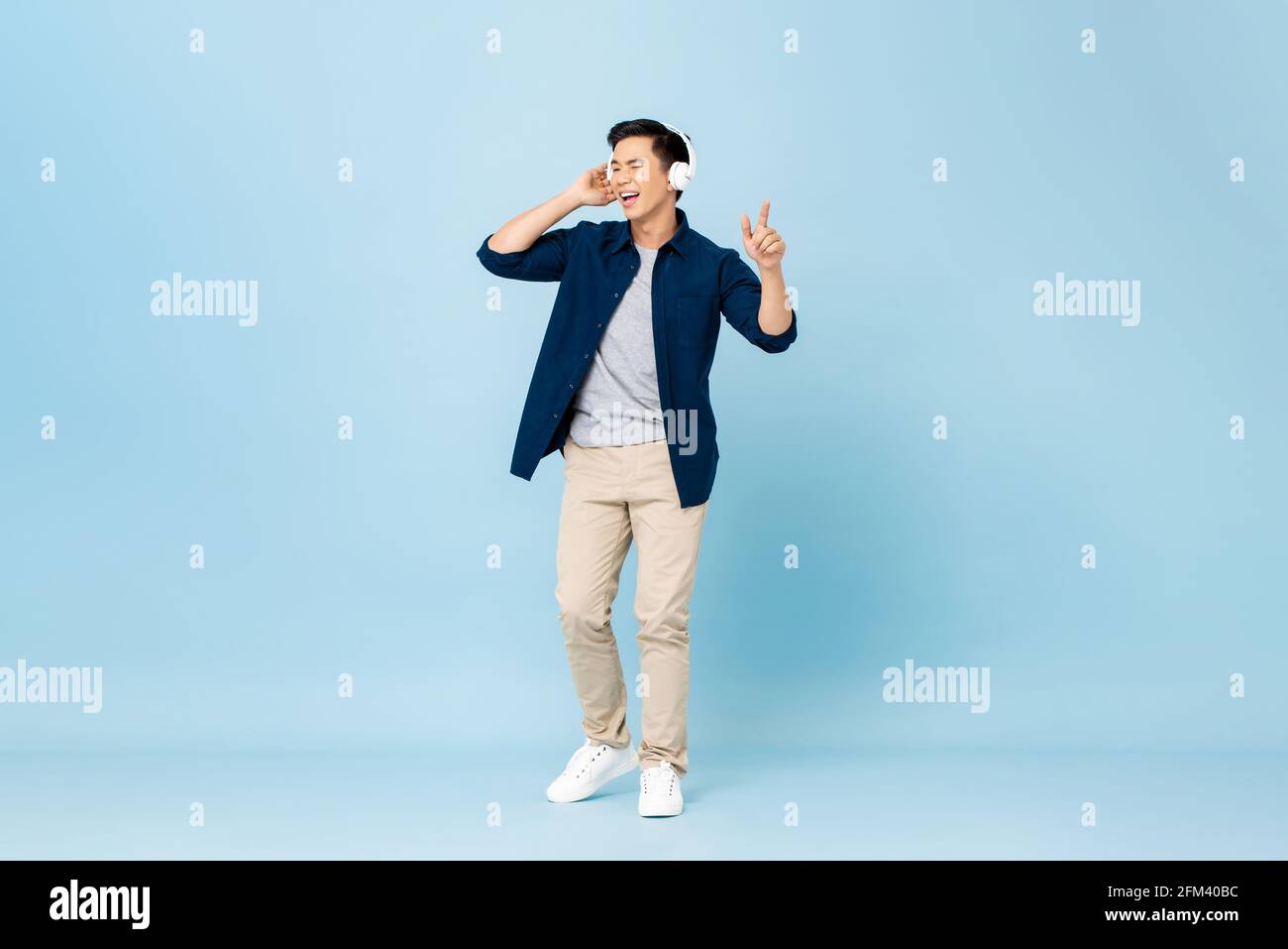 Lächelnder junger, hübscher asiatischer Mann, der Musik streamt Kabellose Kopfhörer im isolierten hellblauen Studiohintergrund Stockfoto