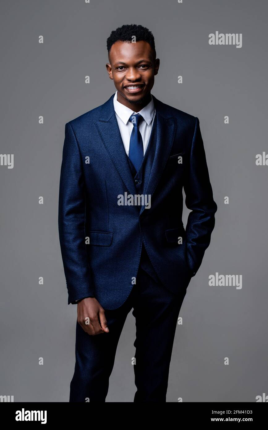 Lächelnder junger, hübscher afrikanischer Mann, der einen formellen Geschäftsanzug im Stehen trägt Mit Hand in Tasche auf Studio grau isoliert Hintergrund Stockfoto