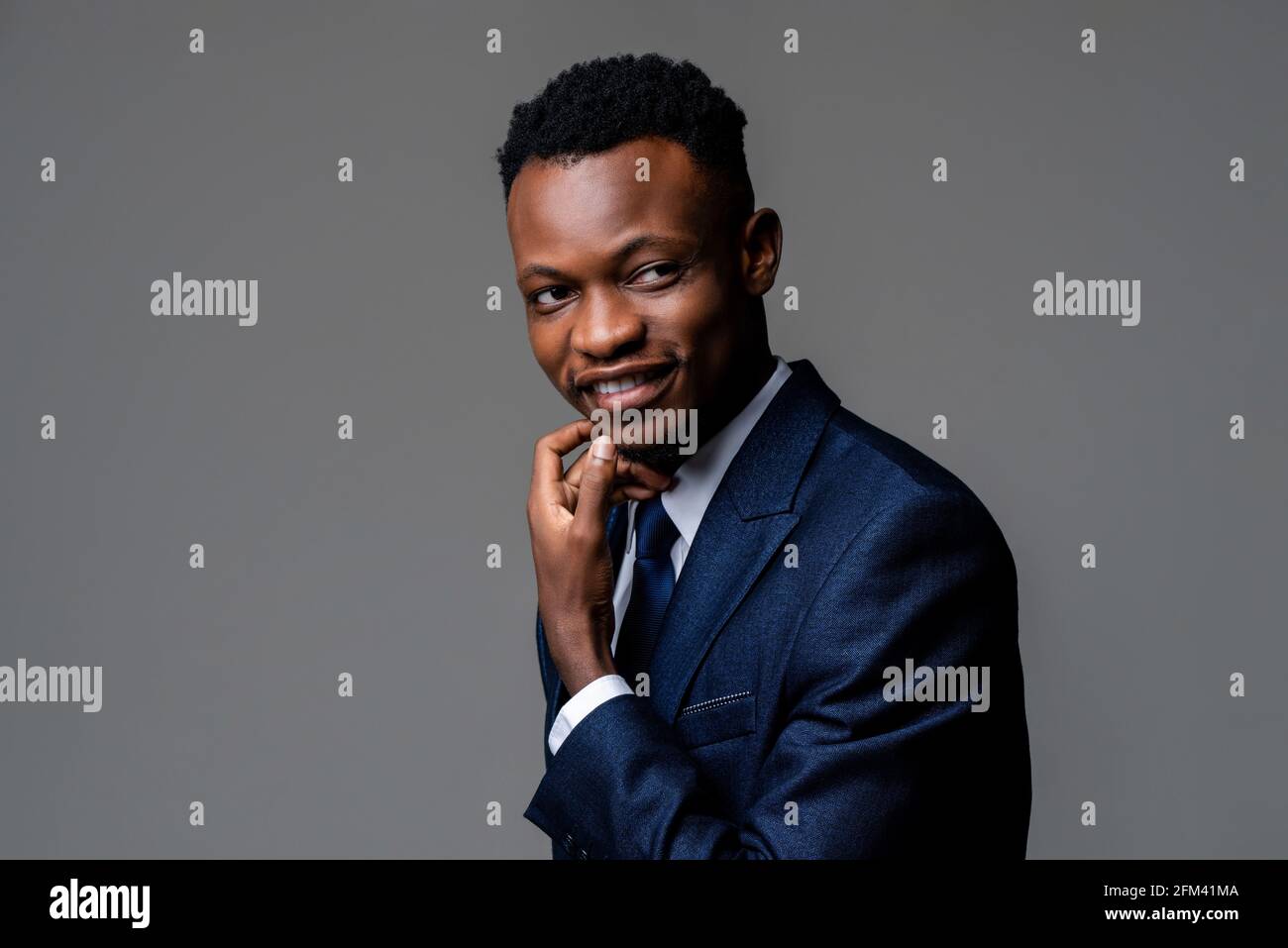Lächelnder junger, hübscher afrikanischer Mann in formellem Geschäftsanzug Mit Hand berühren Gesicht in Studio grau isoliert Hintergrund Stockfoto