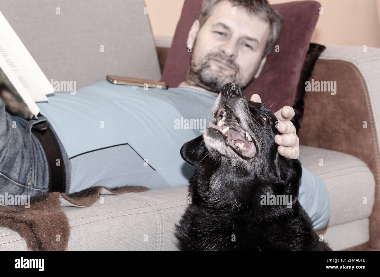 Ein Mann mittleren Alters mit gräulichen Haaren und Bart liegen Auf dem Sofa und streicheln seinen schwarzen älteren Hund Stockfoto