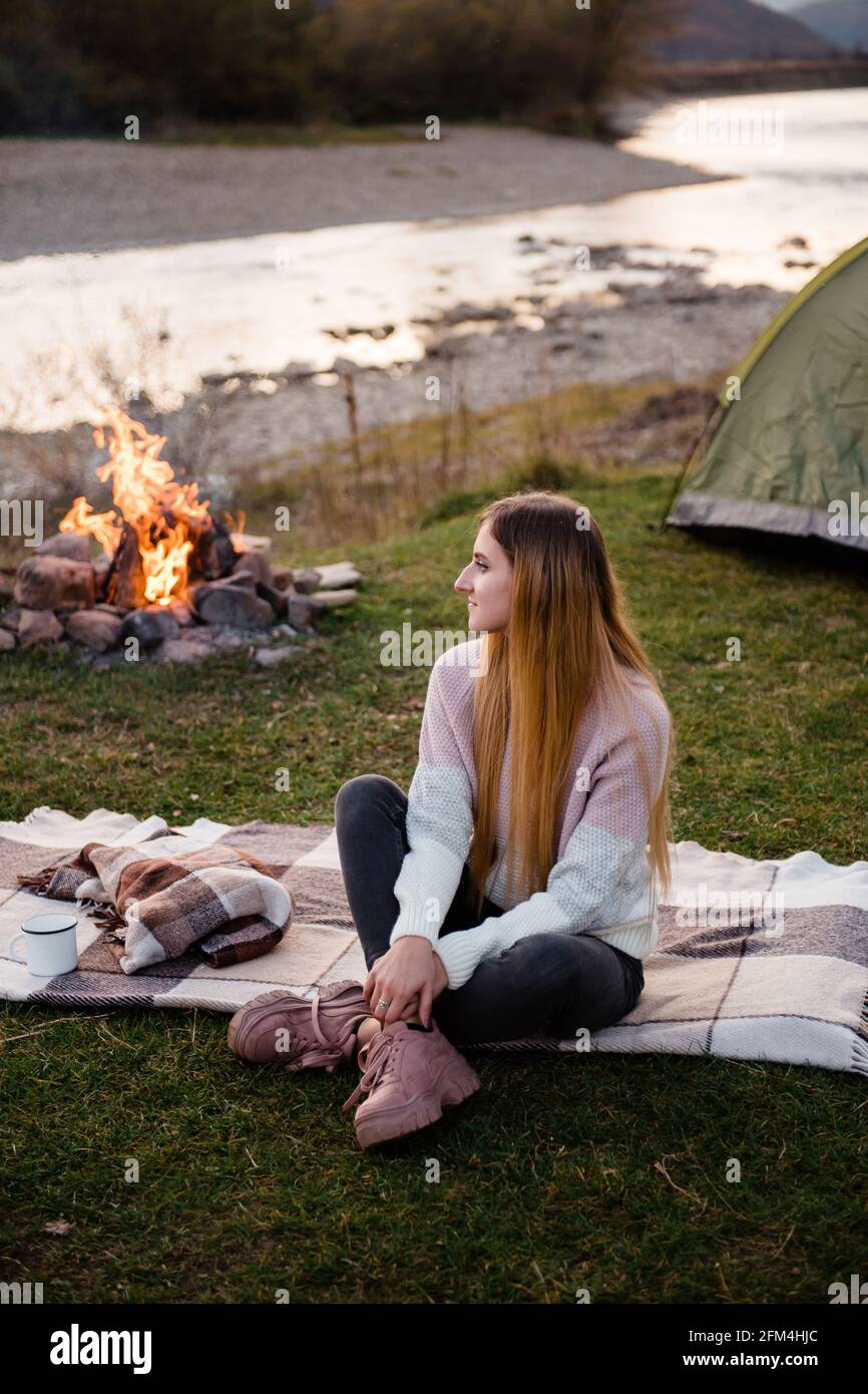 Ruhe in der Nähe der Berge. Das junge Mädchen sitzt am Nachmittag in der Nähe eines Zeltes und genießt den Blick auf die Berge. Das Lagerfeuer ist seitlich beleuchtet. Stockfoto