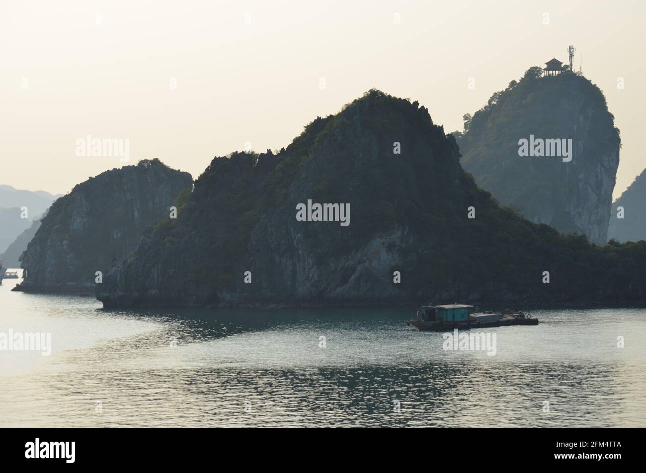 Panoramablick auf den Sonnenuntergang über dem glitzernden Wasser, den Felsformationen, den blauen Bergen und den schwimmenden Booten in der Halong-Bucht in Vietnam Stockfoto