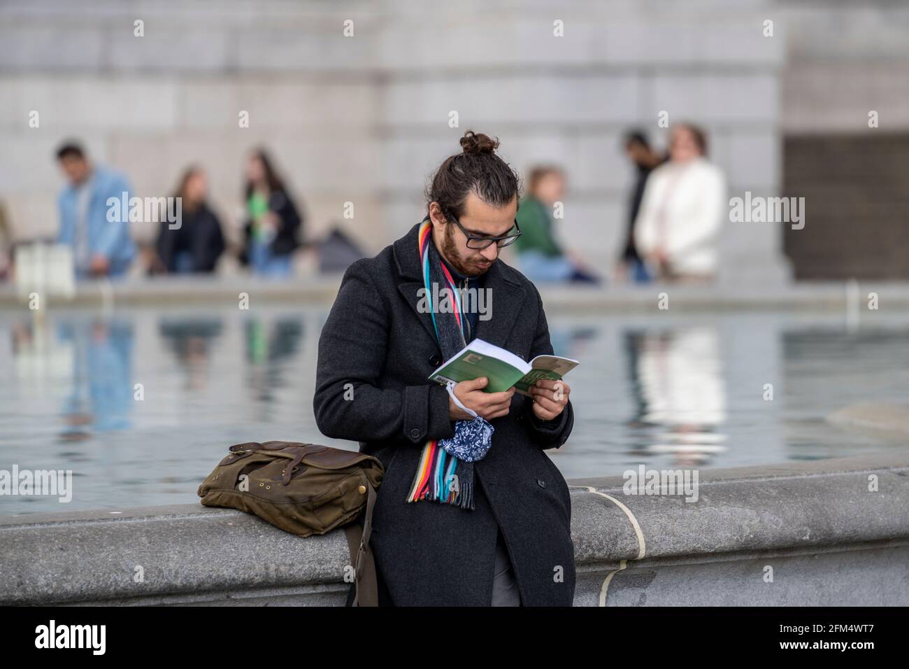 Offen im öffentlichen Park. Ein Mann der Generation z wird gesehen, wie er ein Buch am Rande eines Wasserbrunnens liest. Mit schwarzem Haar in einem Brötchen und einer Brille gebunden. Stockfoto