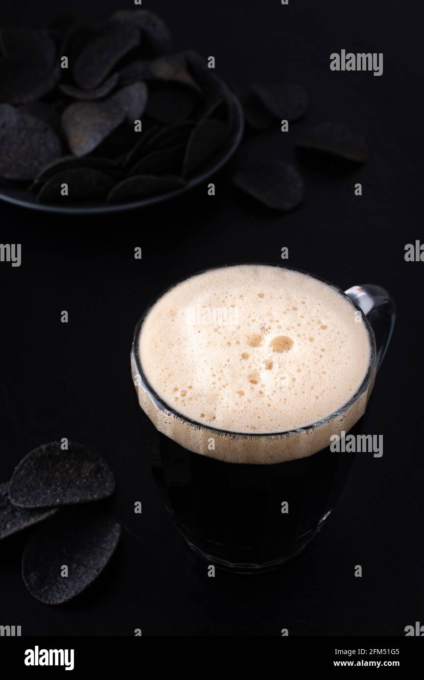 Dunkles Craft-Bier mit Schaum in einem Becher mit Schwarz Kartoffelchips auf dunklem Hintergrund Stockfoto