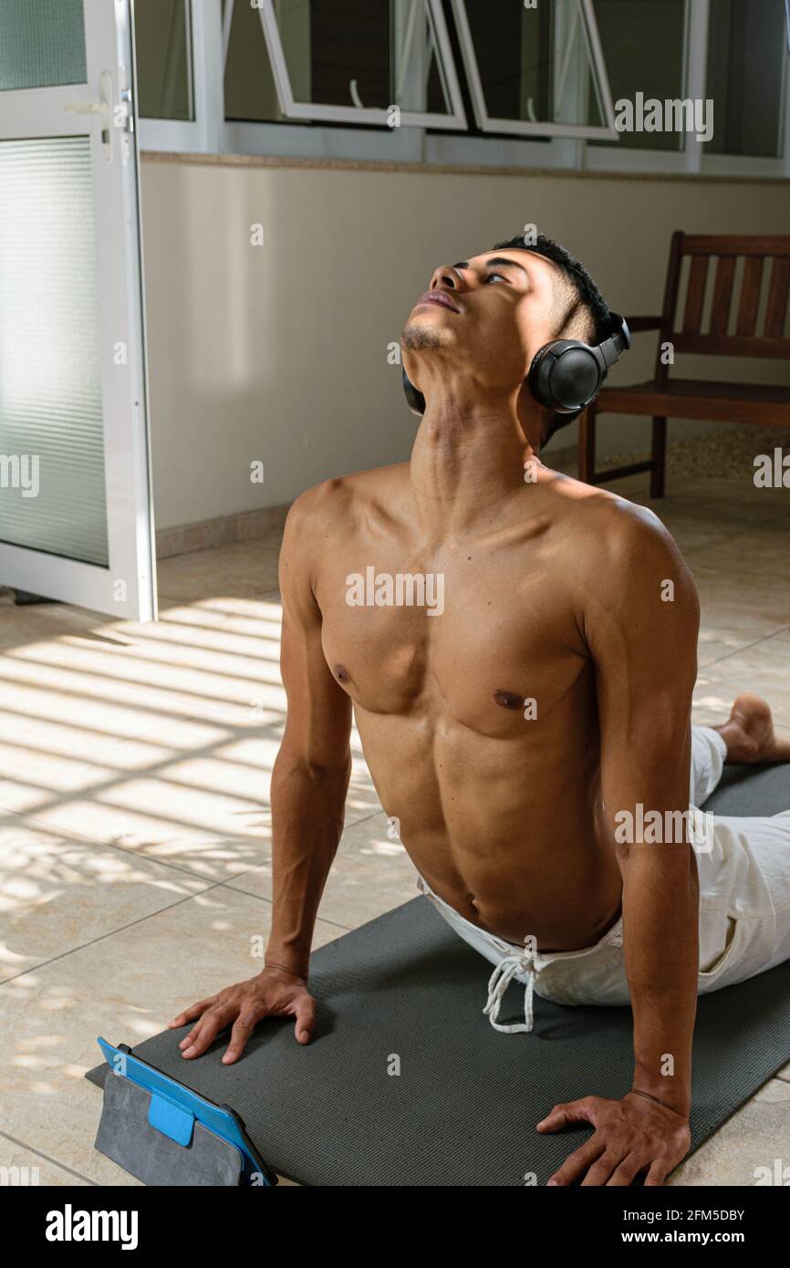 Junger brasilianischer Mann, der mit dem Kopftelefon einen Online-Yoga-Kurs macht, in der nach oben gerichteten Hundeposition, Vorderansicht. Stockfoto
