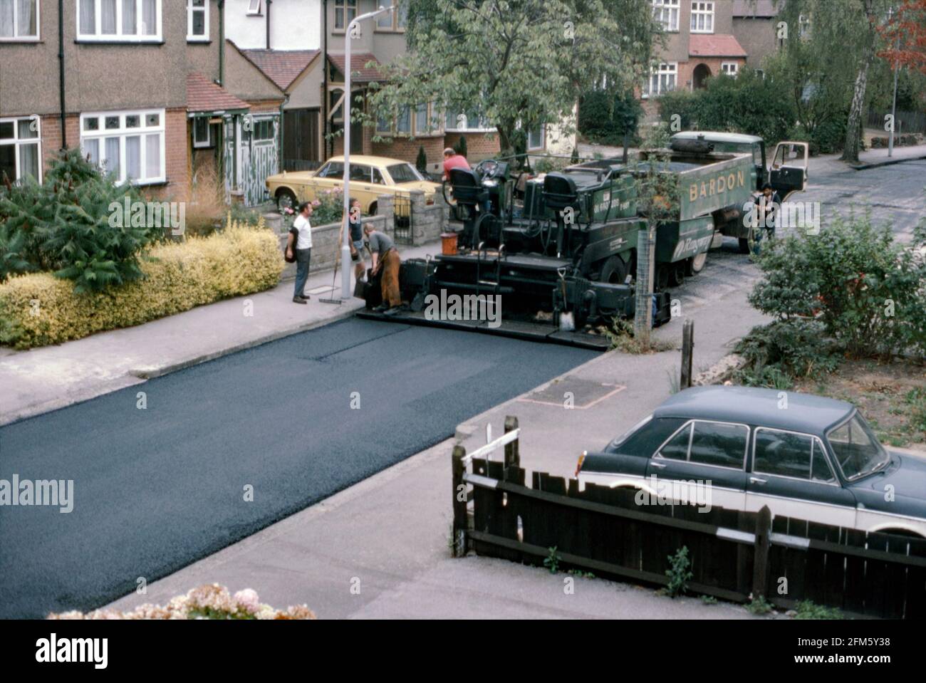 Eine asphaltierte Maschine und Arbeiter, die 1984 eine Vorstadtstraße in London, England, wiederbefahren haben. Ein LKW von der Firma Bardon hat eine Charge frischen Materials an das asphaltierte Legefahrzeug geliefert. Die neue Oberfläche zeigt sich hinter der Maschine. Die Straße ist typisch für Londons äußere ‘Metroland’ Vororte mit Häusern, die größtenteils Doppelhaushälfte sind und aus den 1930er/40er Jahren stammen. Dieses Bild stammt aus einem alten 35-mm-Kodak-Amateurfoto mit Farbtransparenz – einem Vintage-Foto aus den 1980er Jahren. Stockfoto