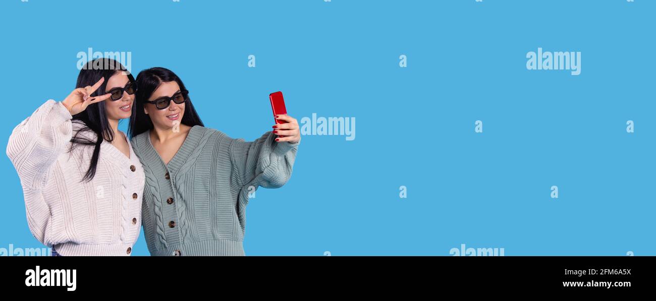 Banner, Langformat. Junge Frauen Zwillinge posieren in einer 3D-Brille und machen ein Selfie auf einem Smartphone. Isoliert auf blauem Studiohintergrund mit leerem Platz für Stockfoto