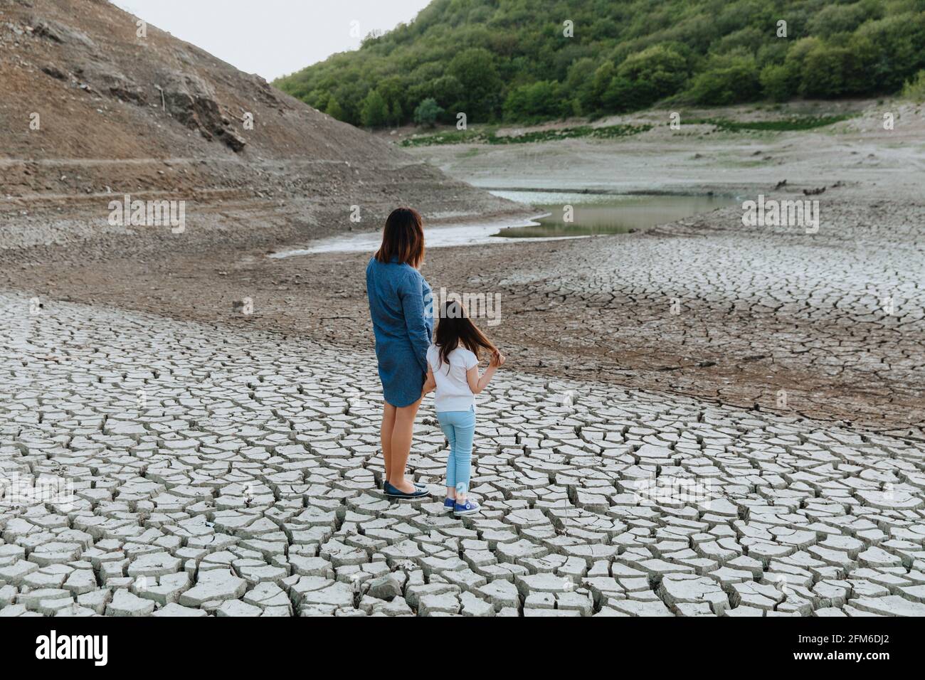 Eine Frau und ein Mädchen stehen Hand in Hand auf trockenem, rissem Boden und schauen auf den See, der austrocknet. Globale Erwärmung. Stockfoto