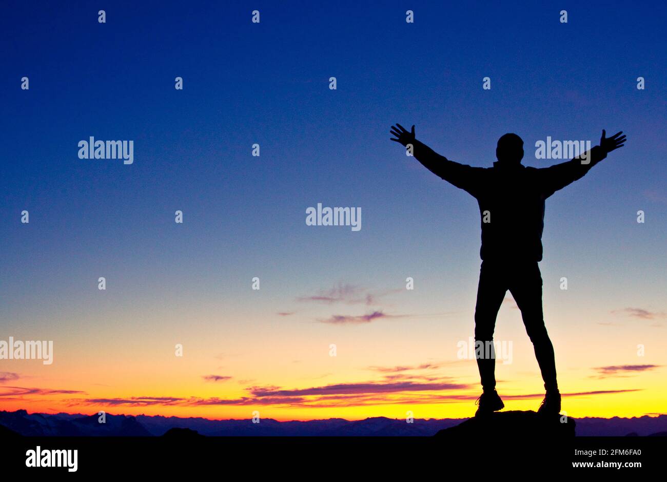 Silhouettierter Wanderer, der mit erhobenen Armen auf dem Berggipfel steht Stockfoto