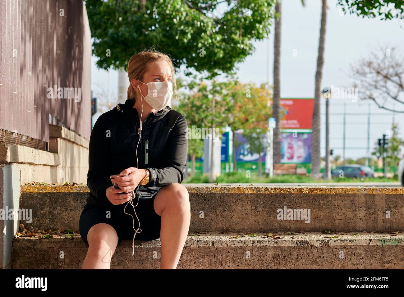 Frau in Sportkleidung und Gesichtsmaske schaut auf ihr Telefon Sitzen auf Treppen Stockfoto