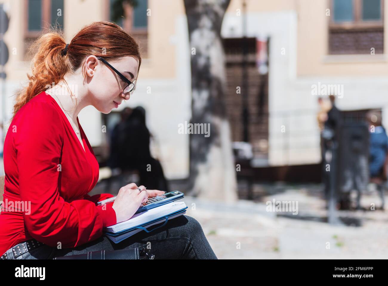 Junge rothaarige Frau mit Brille, die auf einer Bank sitzt, während sie einen Taschenrechner benutzt und etwas in ihr Notizbuch schreibt. In einer roten Bluse gekleidet. Stockfoto