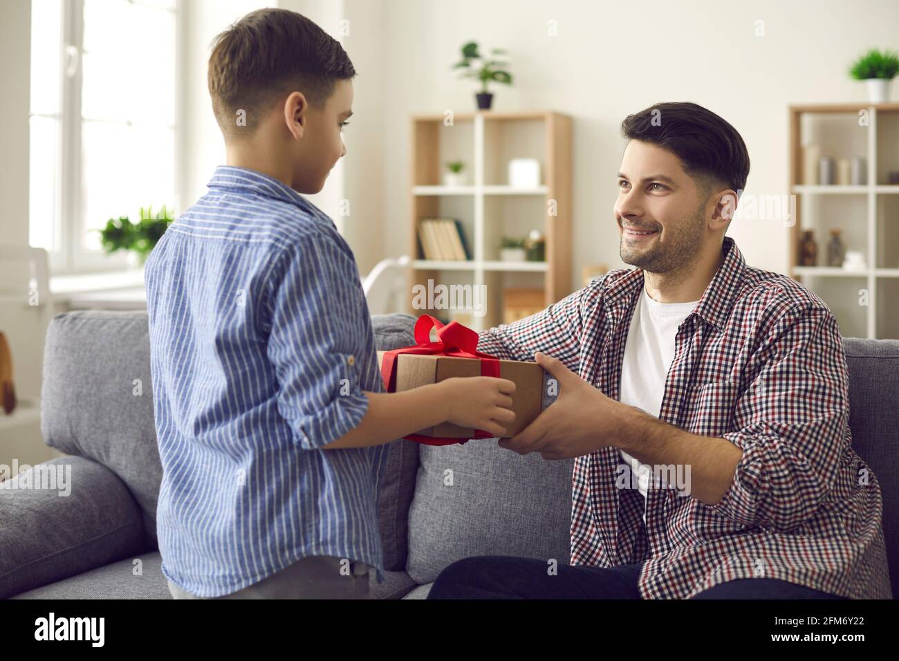 Fröhlicher Vater sitzt auf dem Sofa und nimmt eine Überraschungs-Geschenkbox mit Von den Händen des Sohnes Stockfoto