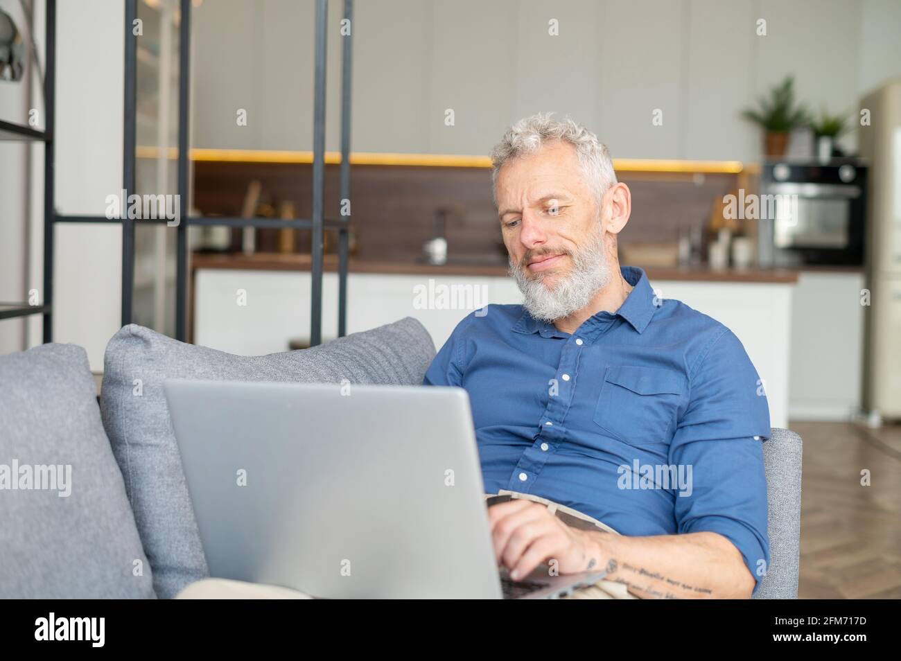 Bärtiger Mann mittleren Alters, der sich mit einem Laptop auf der Couch niederlegt, fernab von zu Hause arbeitet, charismatischer älterer Mann mit lässigem Hemdtippen, E-Mails sendet und in entspannter Atmosphäre zu Hause Nachrichten sendet Stockfoto