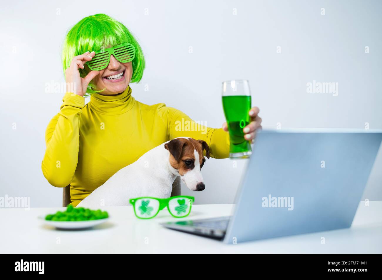 Eine junge Frau in einer grünen Perücke und fröhlichen Gläsern trinkt Bier und beißt glasierte Nüsse. Ein Mädchen sitzt mit einem Hund an einem Tisch und feiert den St. patricks Tag Stockfoto